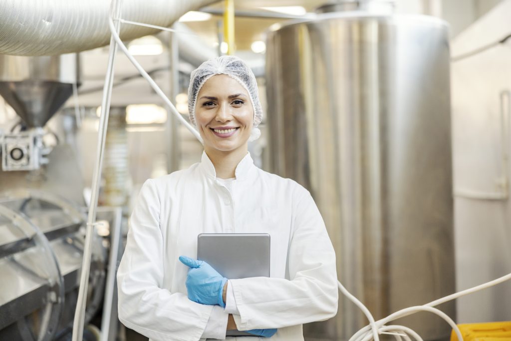 A dairy factory worker is standing in facility and holding tablet while smiling at the camera.