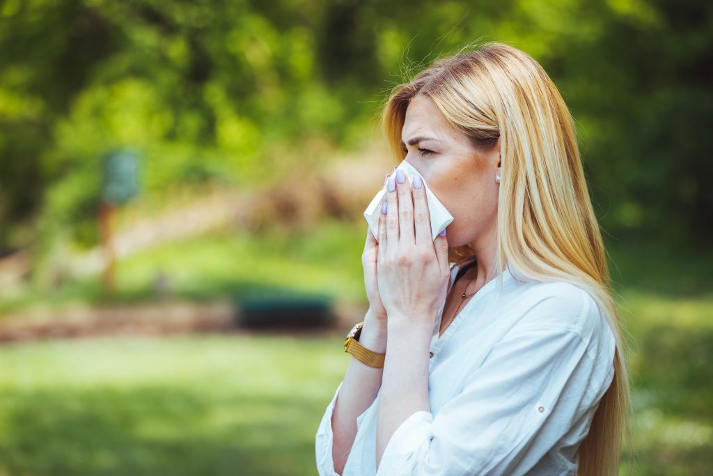 Young pretty woman blowing nose in front of blooming tree. Spring allergy concept. Attractive young adult woman coughing and sneezing outdoors. Sick people allergy or virus influenca concept.