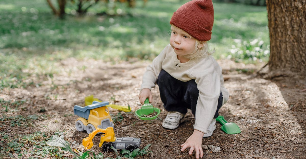 a child wearing a red beanie plays with toy trucks outdoors on a sunny day in a park