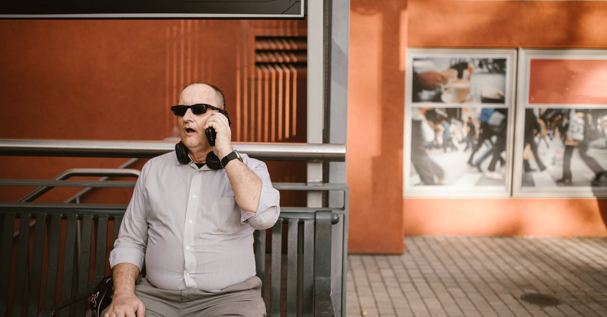 a mature man in sunglasses sits outdoors using a cellphone by a modern building