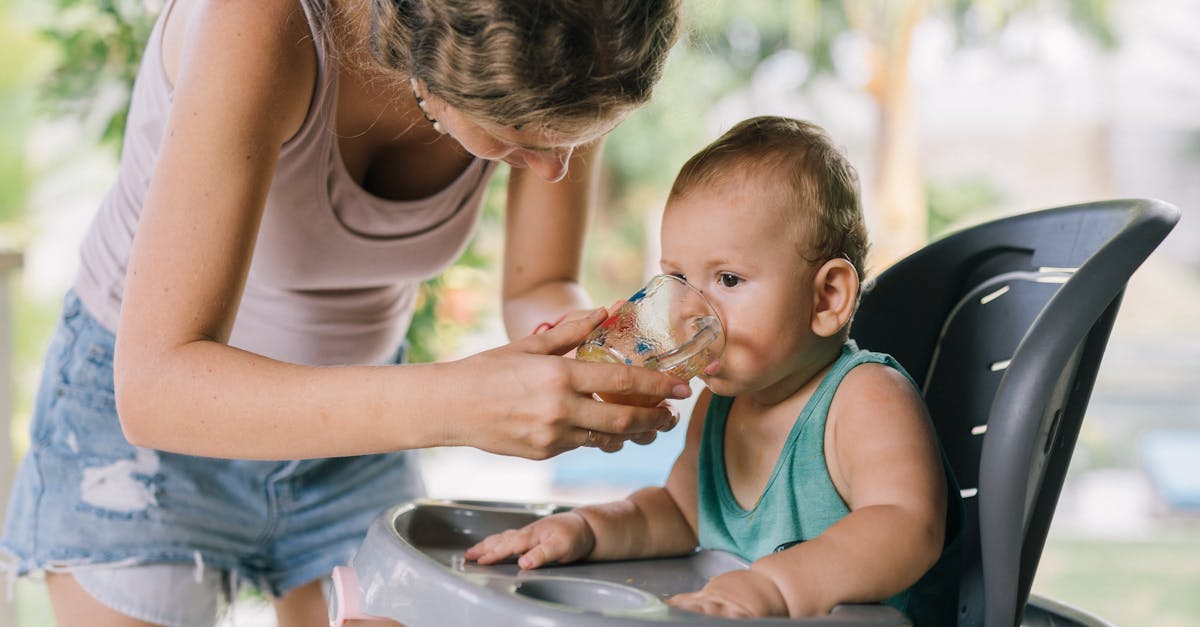 a mother helps her baby drink water from a glass while sitting in a high chair