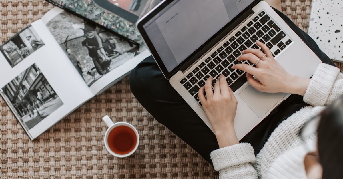 overhead view of a woman typing on a laptop at home surrounded by magazines and a cup of tea on the