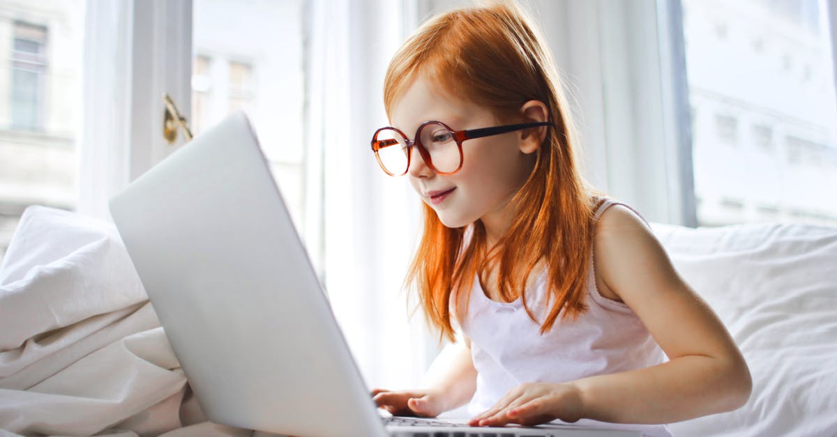 red haired child in glasses using a laptop on a white bed bright room